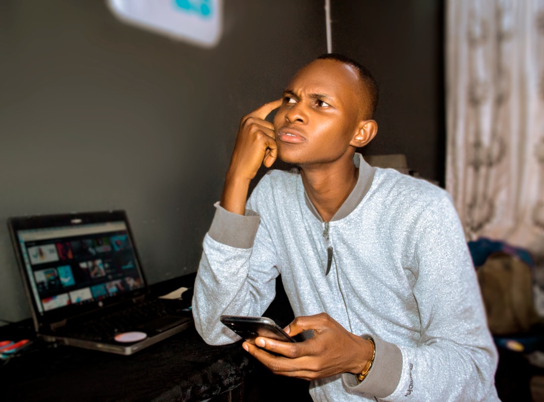 Nigerian business owner looking worried while holding a smartphone at his laptop, contemplating a hiring decision for a digital marketer